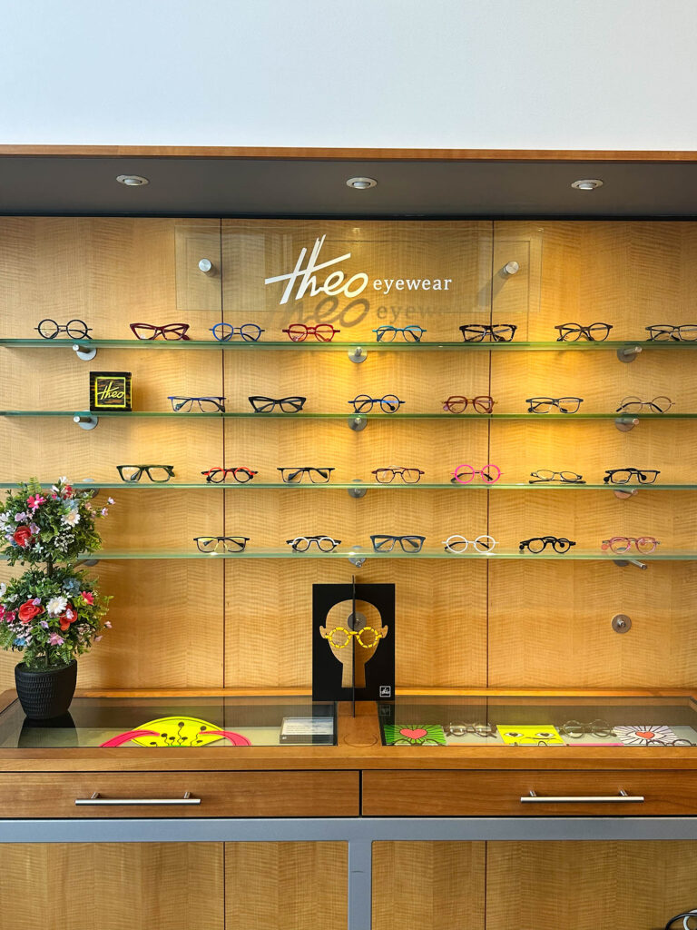 A display of colorful eyewear on glass shelves, featuring various styles and designs. A small sign reads "Theo eyewear," and a potted plant with vibrant flowers sits on a wooden counter below the display.