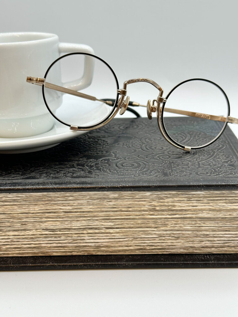 Round eyeglasses with a gold frame rest on a closed, textured book. A white coffee cup sits on a saucer in the background, all placed on a light surface.