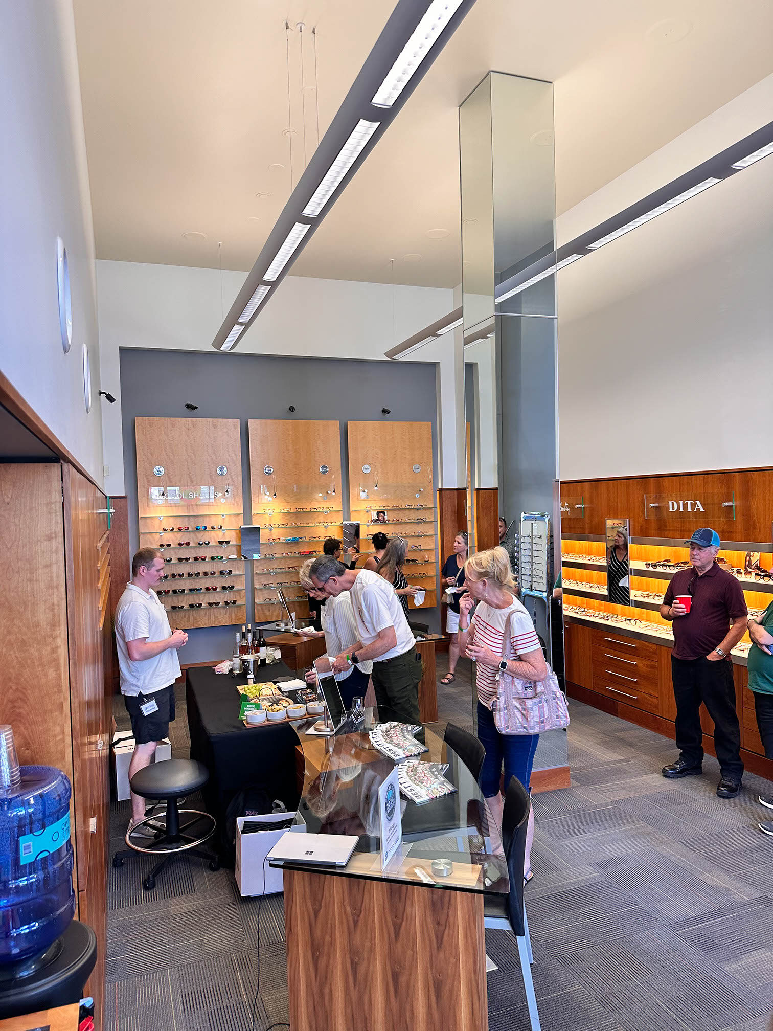 A modern eyewear store with several people browsing. Glasses are displayed on wooden walls, and a sales counter with staff is in the center. Customers are casually dressed, with some engaged in conversation. A water cooler is visible on the left.