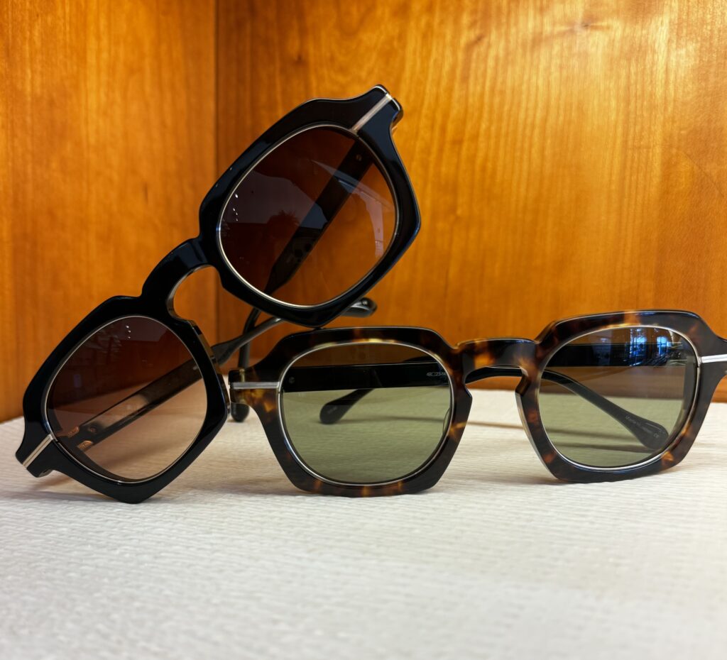 Two pairs of sunglasses are displayed against a wooden background. One pair is black with dark lenses, and the other is tortoiseshell with green-tinted lenses, both resting on a white surface.