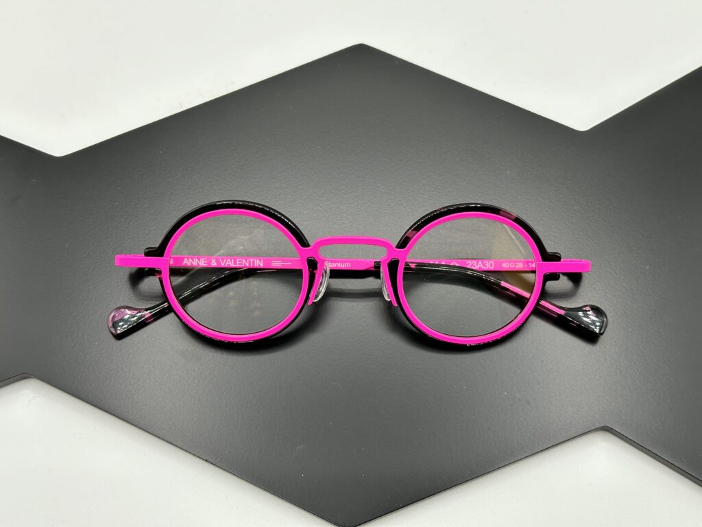 Round eyeglasses with bright pink frames and black accents are centered on a dark, angular surface. The brand name "Anne & Valentin" is visible on the temple arm.