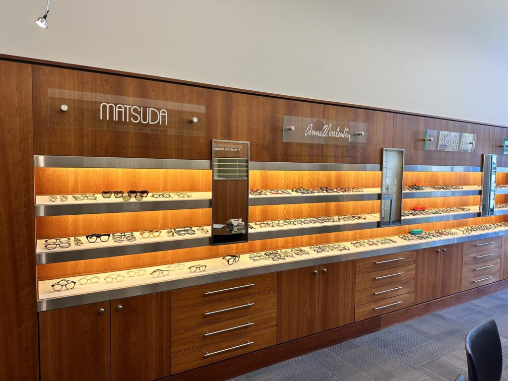 A well-lit eyeglass display in a store showcases various frames on wooden shelves. Brand names like Matsuda and others are visible above the shelves. The floor is carpeted, and the overall setting appears modern and organized.