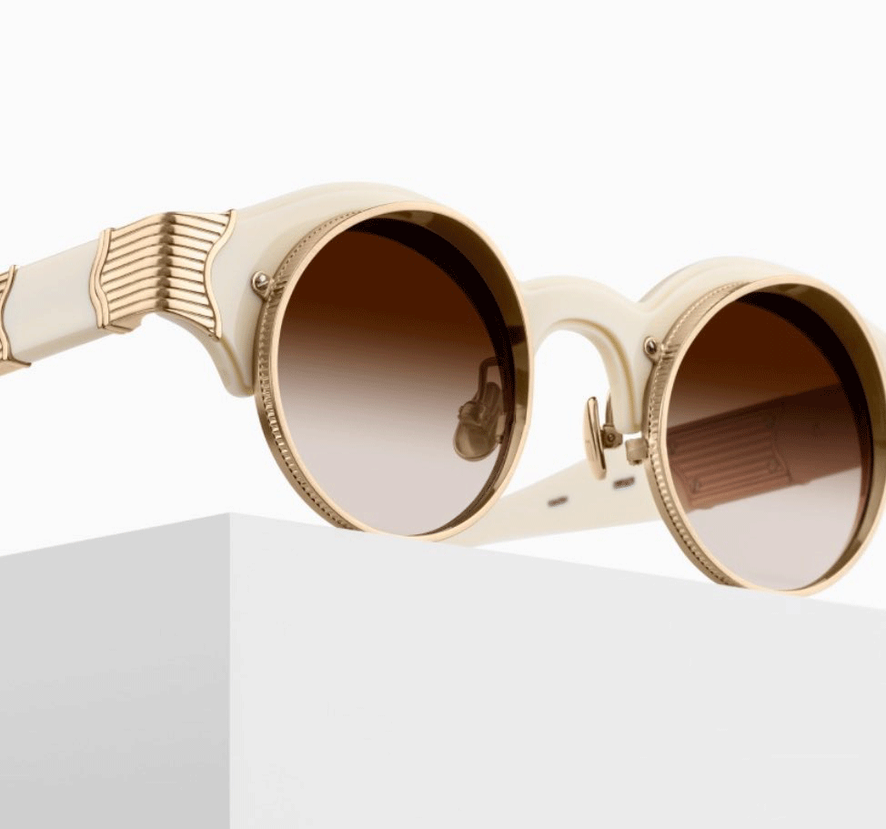 A pair of round sunglasses with cream-colored frames and gold accents rest on a white surface. The lenses are a gradient brown. The design features intricate detailing on the arms and bridge, creating a vintage-inspired look.