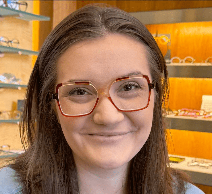 A person with long brown hair is wearing oversize, square-shaped glasses in a store. Shelves with various glasses are blurred in the background. The person is smiling warmly at the camera.