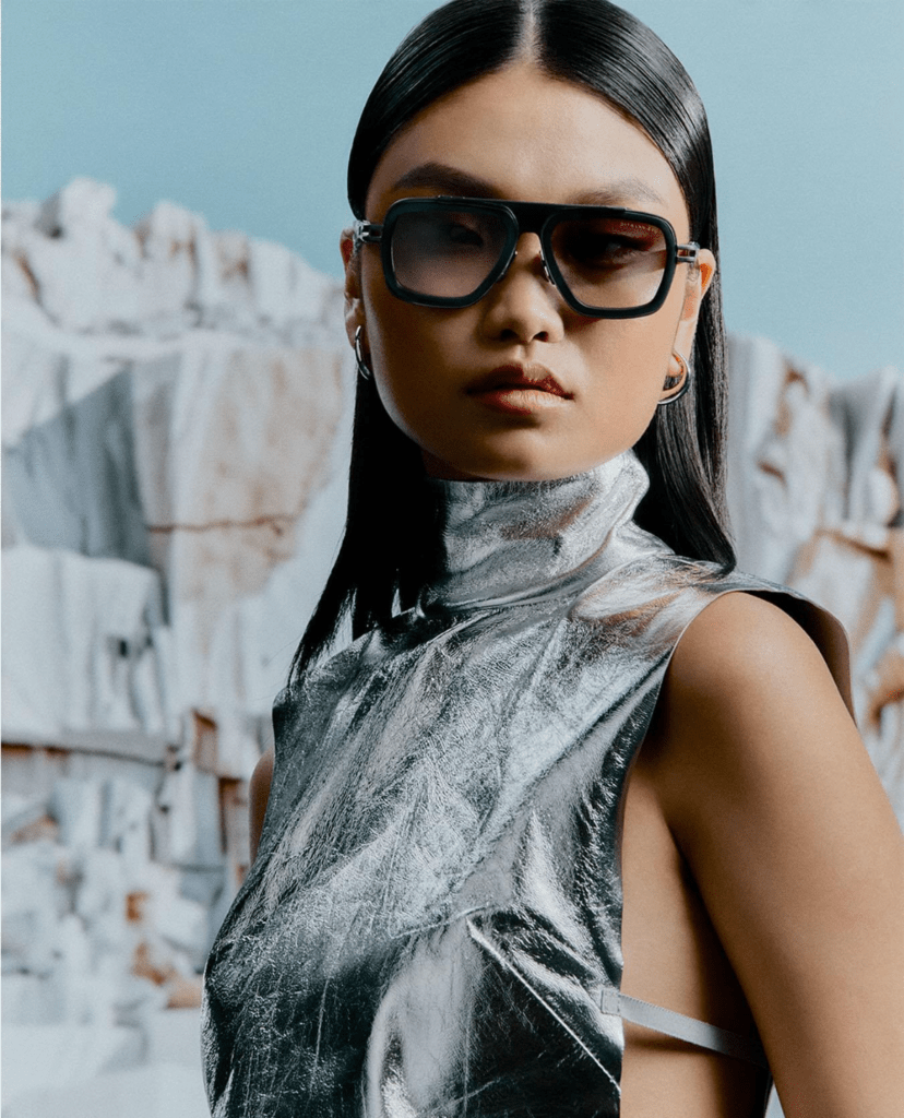 A woman wearing large sunglasses and a metallic silver sleeveless top stands against a backdrop of rocky cliffs under a pale blue sky. Her hair is straight and parted in the middle.