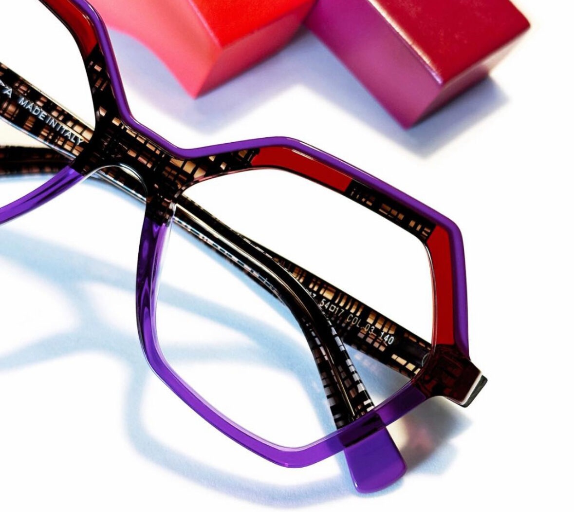 Close-up of geometric purple eyeglass frames with intricate detailing on the temples, resting on a white surface. In the background, blurred red and orange rectangular objects are partially visible.