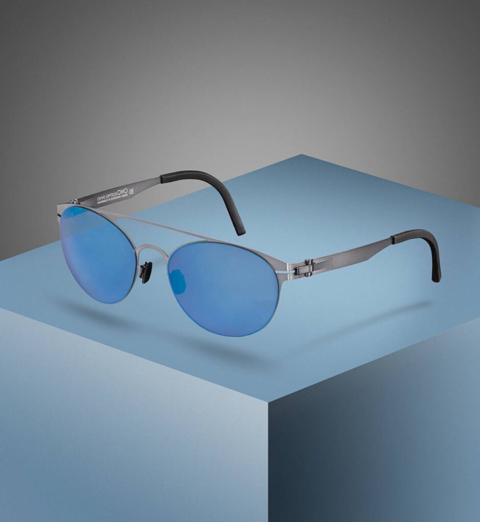 Sleek, silver-framed sunglasses with blue-tinted lenses displayed on a minimalist blue and gray geometric pedestal against a gray gradient background.