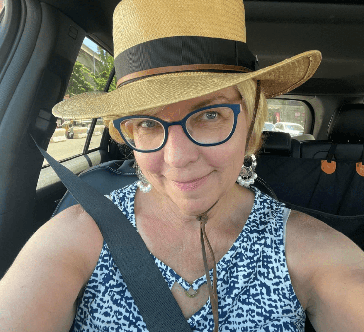 A person wearing a straw hat and blue-rimmed glasses is smiling while sitting in a car. They are wearing a sleeveless blue and white patterned top and a seatbelt is fastened. Sunlight is coming through the window.