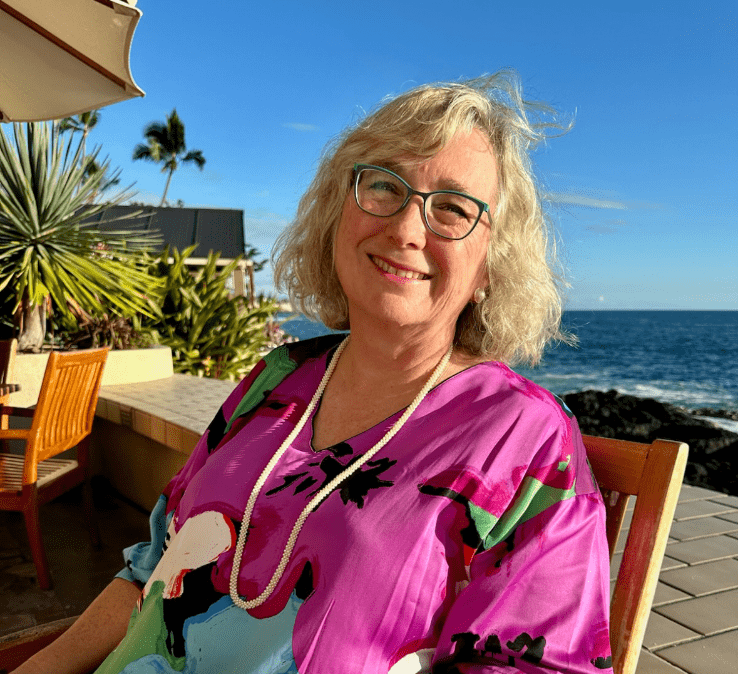 A smiling woman with glasses and curly blonde hair sits at an outdoor table. She's wearing a colorful blouse and necklace, with the ocean and palm trees in the background under a clear blue sky.