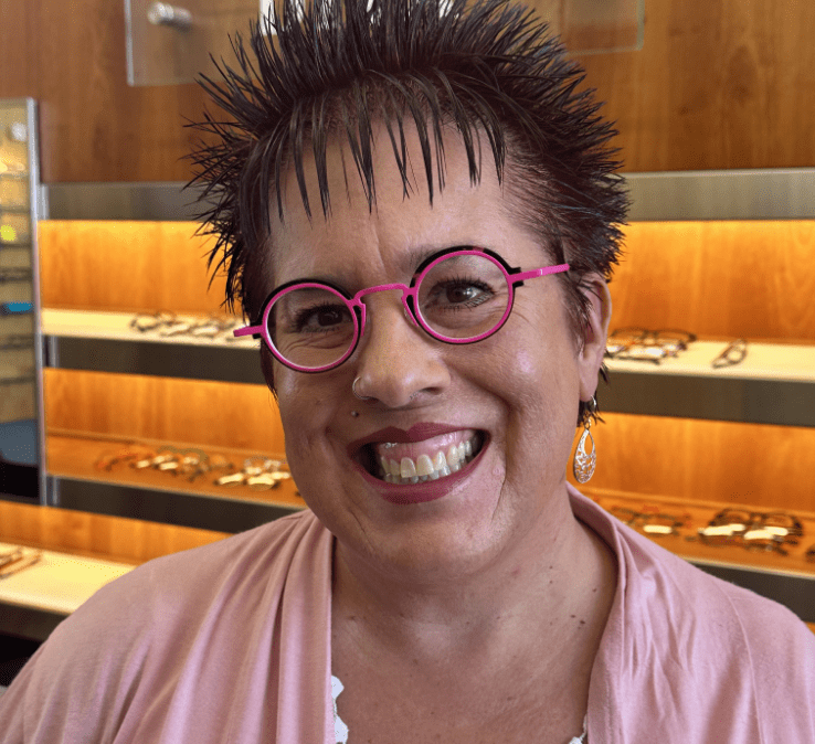 A person with short, spiky hair smiles while wearing pink glasses. They're dressed in a pink top and standing in front of wooden shelving displaying various eyeglasses.