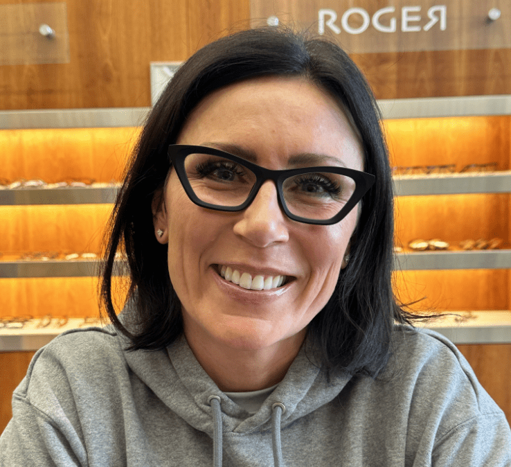 A woman with shoulder-length dark hair and cat-eye glasses smiles warmly. She is wearing a gray hoodie. Behind her are wooden shelves with various items displayed on them.