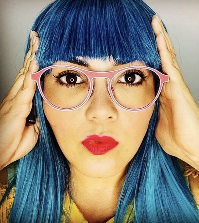 Person with long blue hair and bangs wearing pink glasses and red lipstick, holding their hands to the sides of their head, looking directly at the camera with a neutral expression.