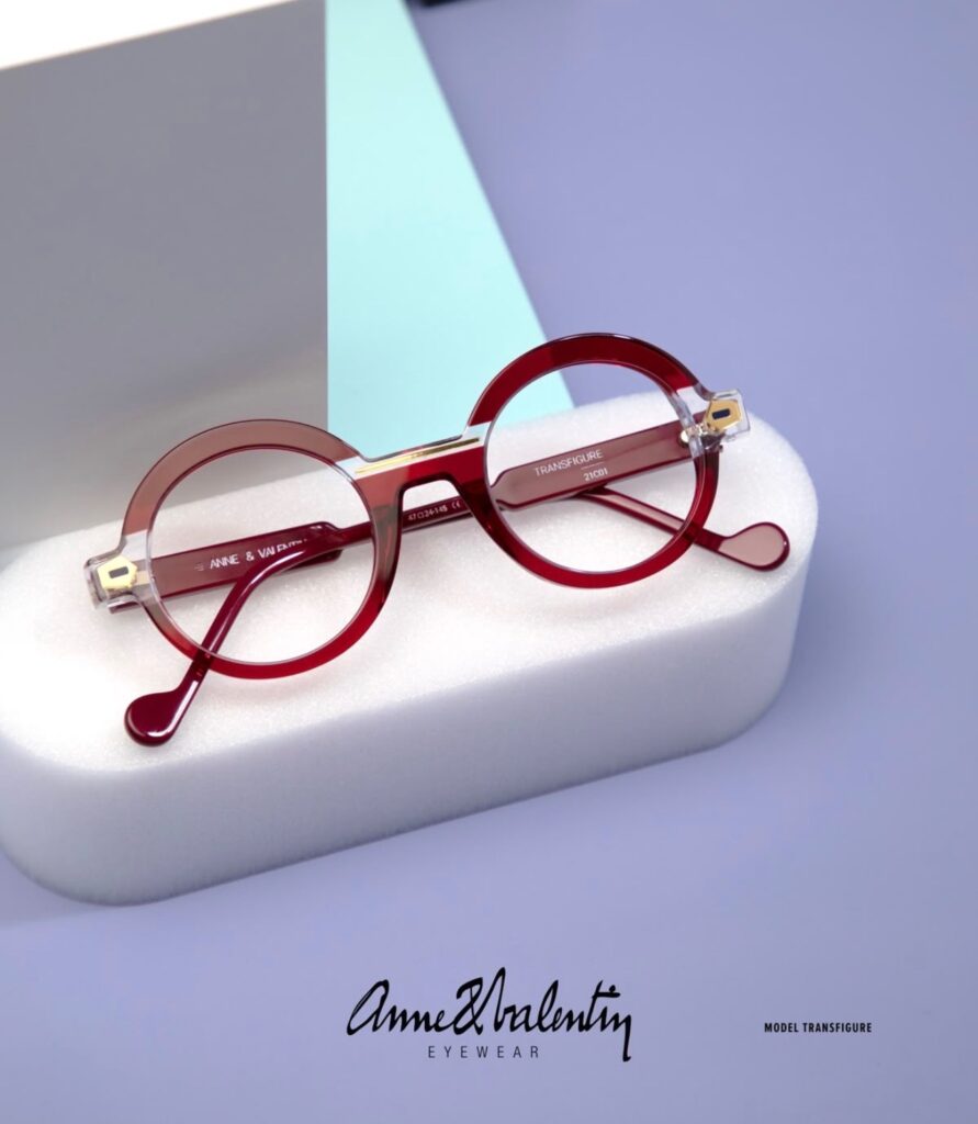 Round, red eyeglasses with thick frames rest on a white foam block against a light purple background. The words "Anne & Valentin EYEWEAR" and "MODEL TRANSFIGURE" are printed at the bottom.