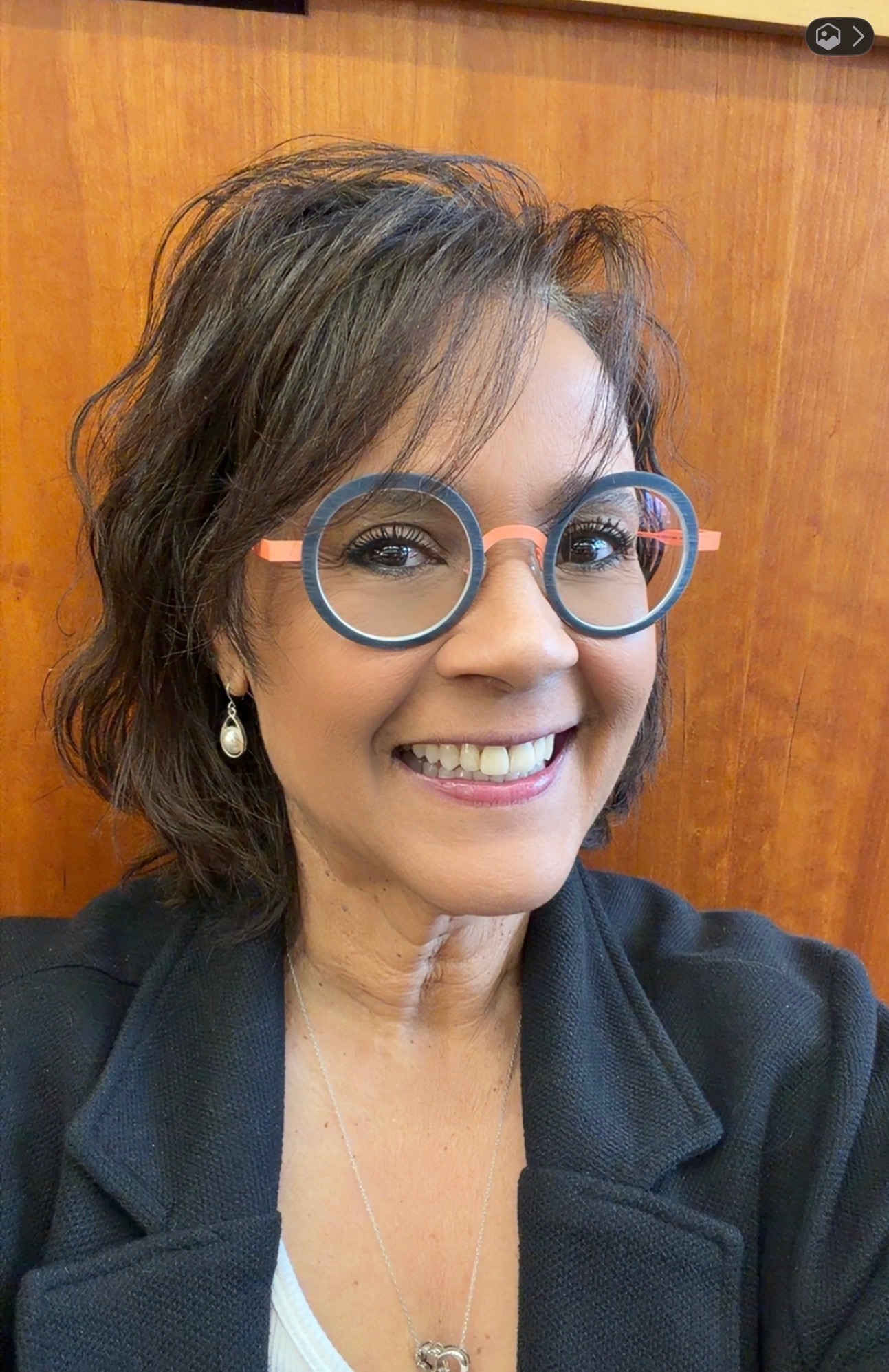 A smiling woman with short dark hair wears round, colorful glasses and a black blazer, posing in front of a wooden background. She has pearl drop earrings and a necklace.