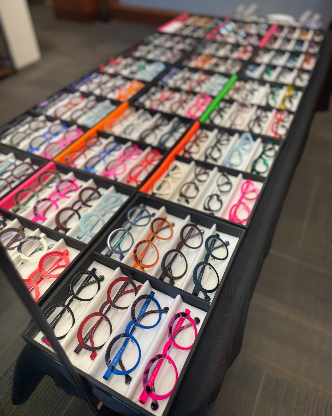 A display table filled with various colorful eyeglass frames organized in trays, featuring a wide range of styles and colors, set on a dark cloth in a showroom or store.
