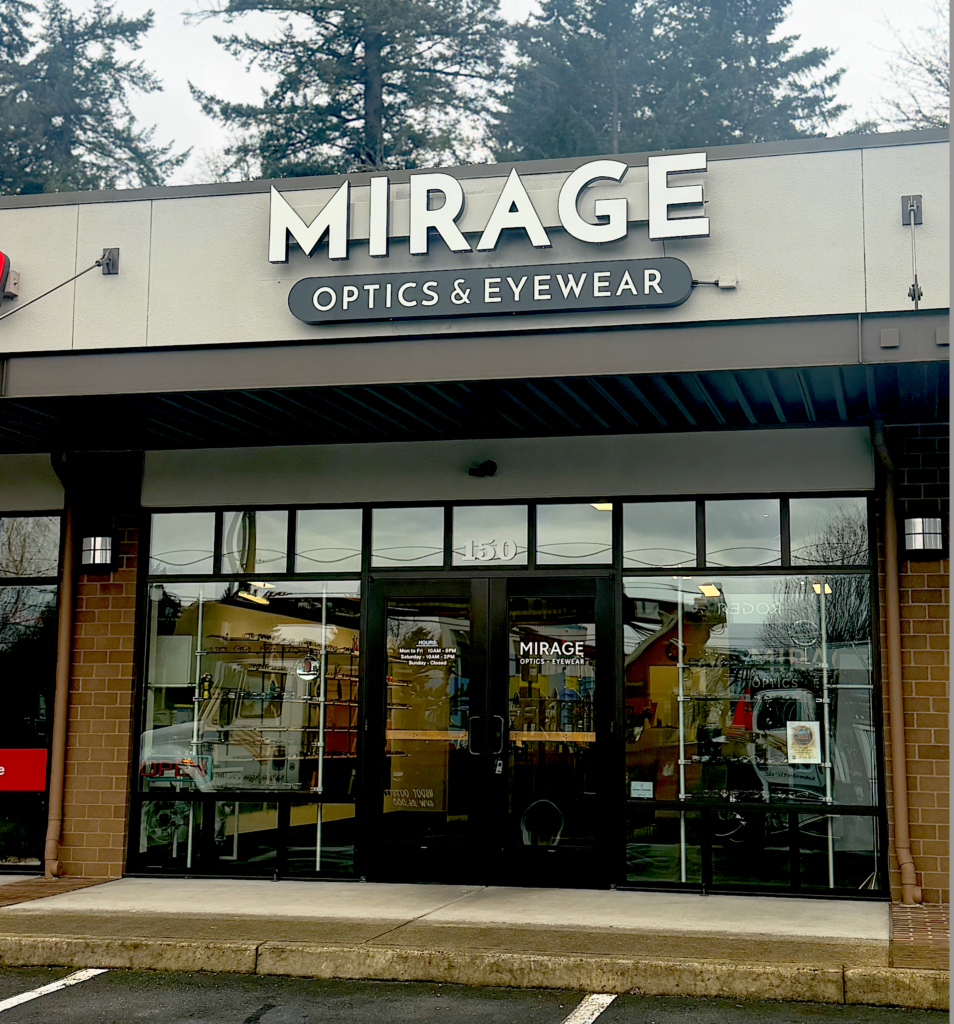 Storefront of “Mirage Optics & Eyewear” with large glass windows and door, displaying eyewear inside. The store is in a strip mall, with trees and a cloudy sky in the background.