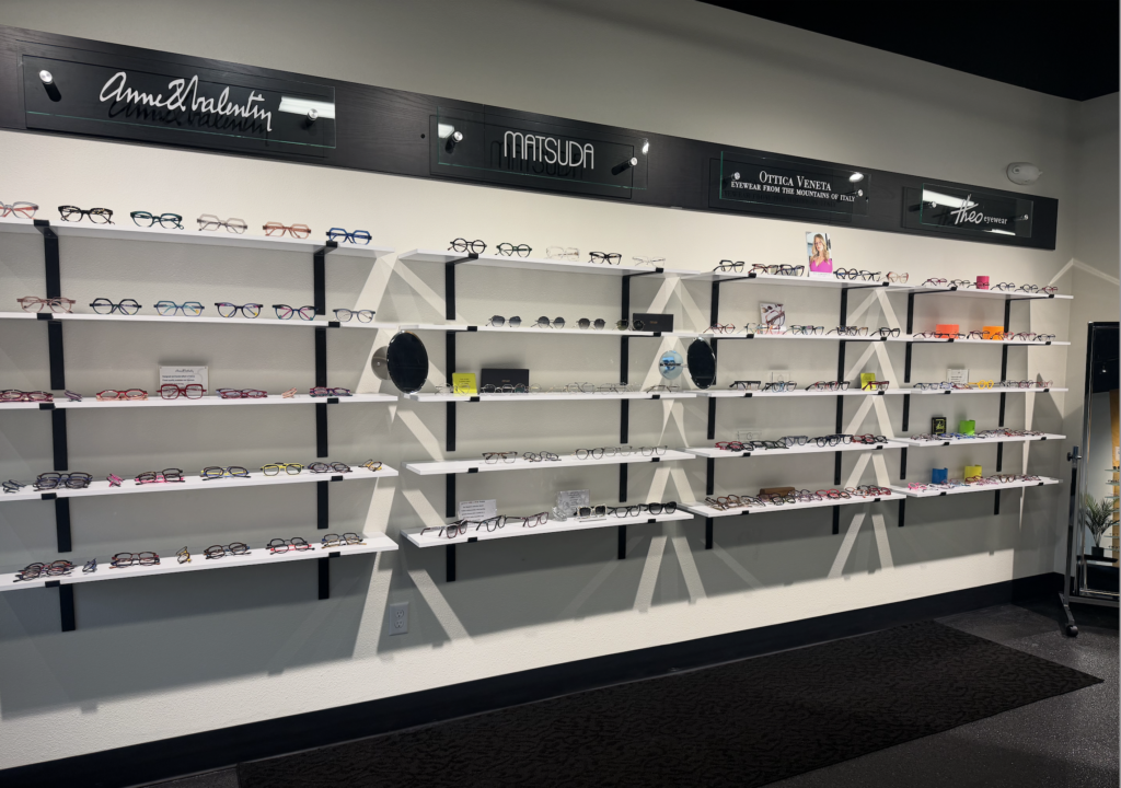 A wall display in an eyewear store showcases various eyeglass frames arranged neatly on multiple white shelves, with brand signs like Anne & Valentin, Matsuda, and Optika Venezia above.