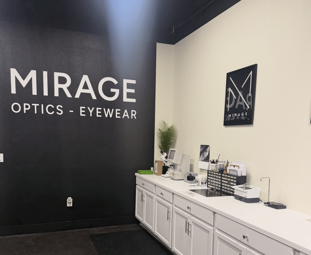 A modern eyewear store interior featuring a black wall with "MIRAGE OPTICS - EYEWEAR" in large white letters, white cabinets, office supplies, and a decorative plant on the countertop.