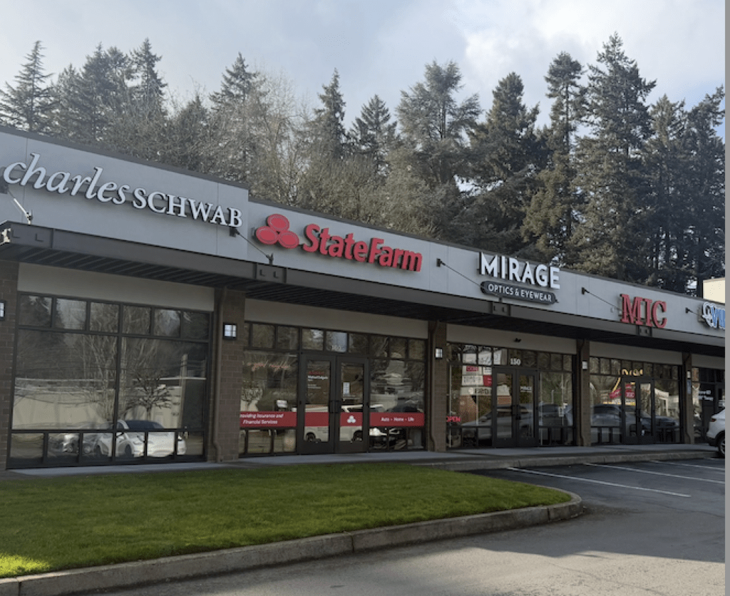 A strip mall with storefronts for Charles Schwab, State Farm, Mirage Optics & Eyewear, and MIC. The buildings are surrounded by trees and the parking lot is in front.